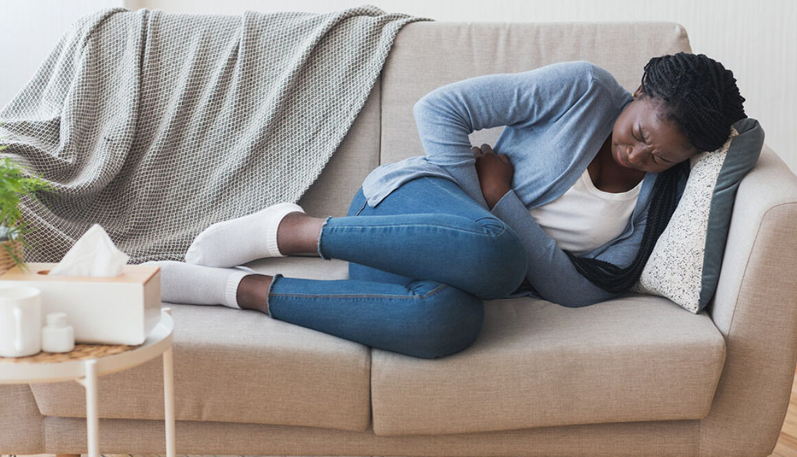 A Young African American Woman Lying on the Couch Clutching Her Stomach in Pain Fibroids vs Cysts