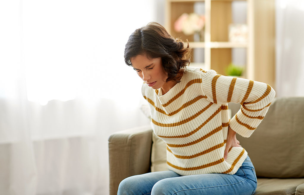 A Woman Sitting On A Couch Grabbing Her Lower Back In Pain Can A UTI Cause Back Pain