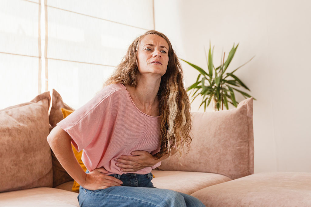 A Young Woman Sitting On A Couch Holding Her Lower Abdomen In Discomfort PMS Vs Pregnancy Symptoms